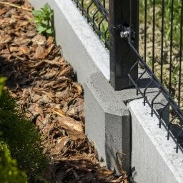 Ogrodzenia Systemowe - Fragment ogrodzenia panelowego z podmurówką betonową, widok z bliska na słupek i mocowanie panela. Kora rozsypana wzdłuż ogrodzenia, zieleń w tle.