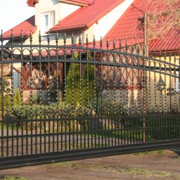 Ogrodzenia Systemowe - Otwarta, czarna, metalowa brama kuta z dekoracyjnymi elementami na tle domu z czerwoną dachówką i kamiennych słupków ogrodzeniowych. Widok z bliska.