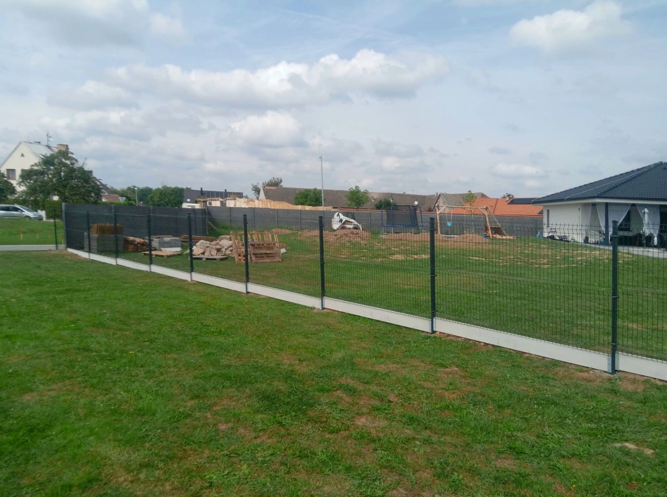 Nowoczesne ogrodzenie panelowe z podmurówką betonową na zielonym trawniku. W tle domy jednorodzinne i błękitne niebo z chmurami. Widok z poziomu gruntu.