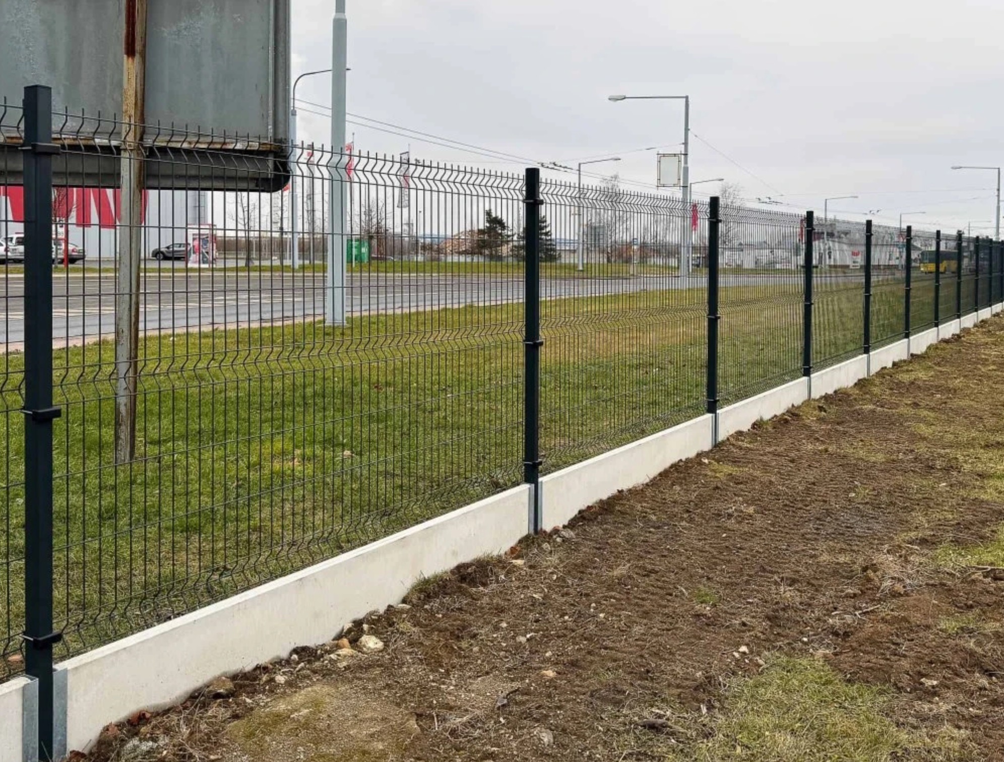 Długi, panelowy płot z podmurówką betonową, ciągnący się wzdłuż drogi. Słupki i panele w kolorze grafitowym. Trawa i ziemia na pierwszym planie, budynki w tle.