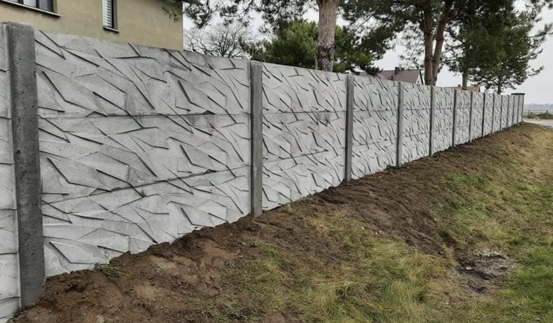 Długi, szary mur z betonu z geometrycznym wzorem, oddzielający posesję od drogi. Widoczne słupki i panele. Trawa i ziemia u podnóża. W tle dom i drzewa.