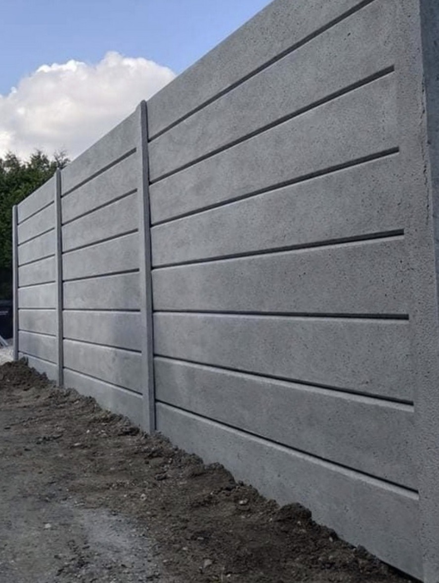 Szary, prefabrykowany mur z betonu architektonicznego, z poziomymi żłobieniami, tworzący nowoczesne ogrodzenie posesji. Widok z dołu, z ziemią u podstawy.