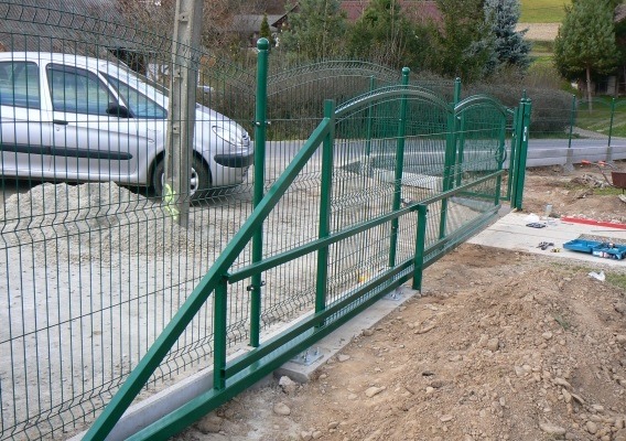 Zielona, przesuwna brama z siatki panelowej, widoczna z boku, z elementami montażowymi i narzędziami na ziemi. W tle zaparkowany samochód i dom.