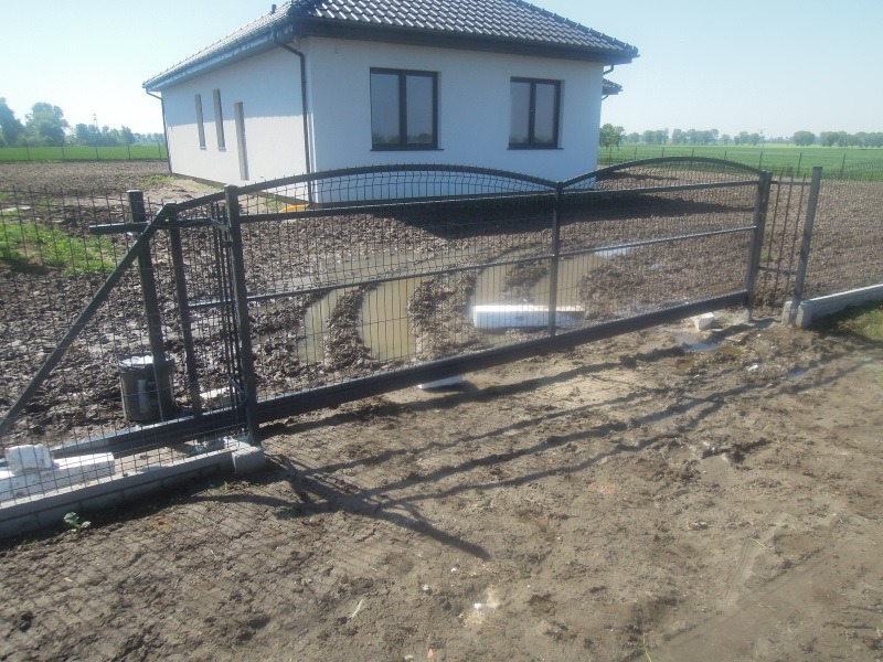 Nowoczesna, czarna brama przesuwna z metalową siatką na tle domu z ciemnym dachem i błotnistego terenu. Widoczne betonowe fundamenty i elementy konstrukcyjne.