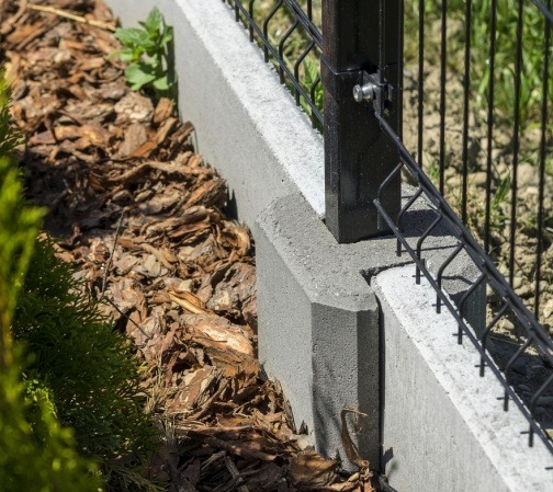 Fragment ogrodzenia panelowego z podmurówką betonową, widok z bliska na słupek i mocowanie panela. Kora rozsypana wzdłuż ogrodzenia, zieleń w tle.