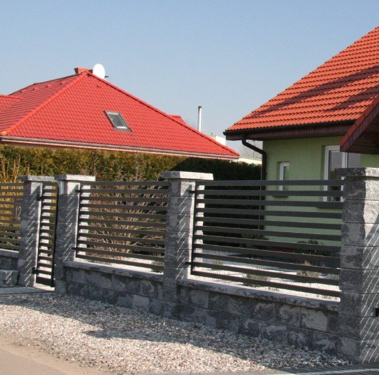 Nowoczesne ogrodzenie z poziomych, grafitowych przęseł i kamiennych słupków, brama wejściowa z numerem 12, na tle domów z czerwonymi dachami.