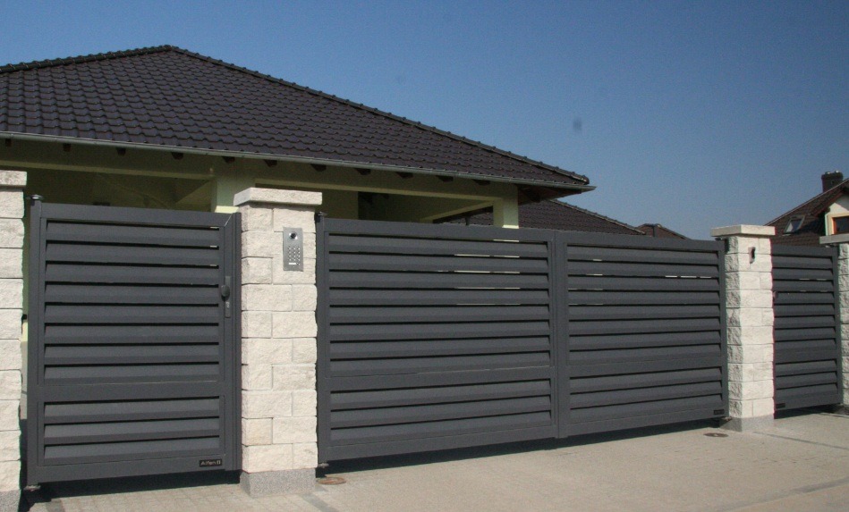 Nowoczesna, przesuwna brama w kolorze antracytu z poziomymi lamelami, osadzona na kamiennych słupkach. Widoczny dom z ciemnym dachem w tle.