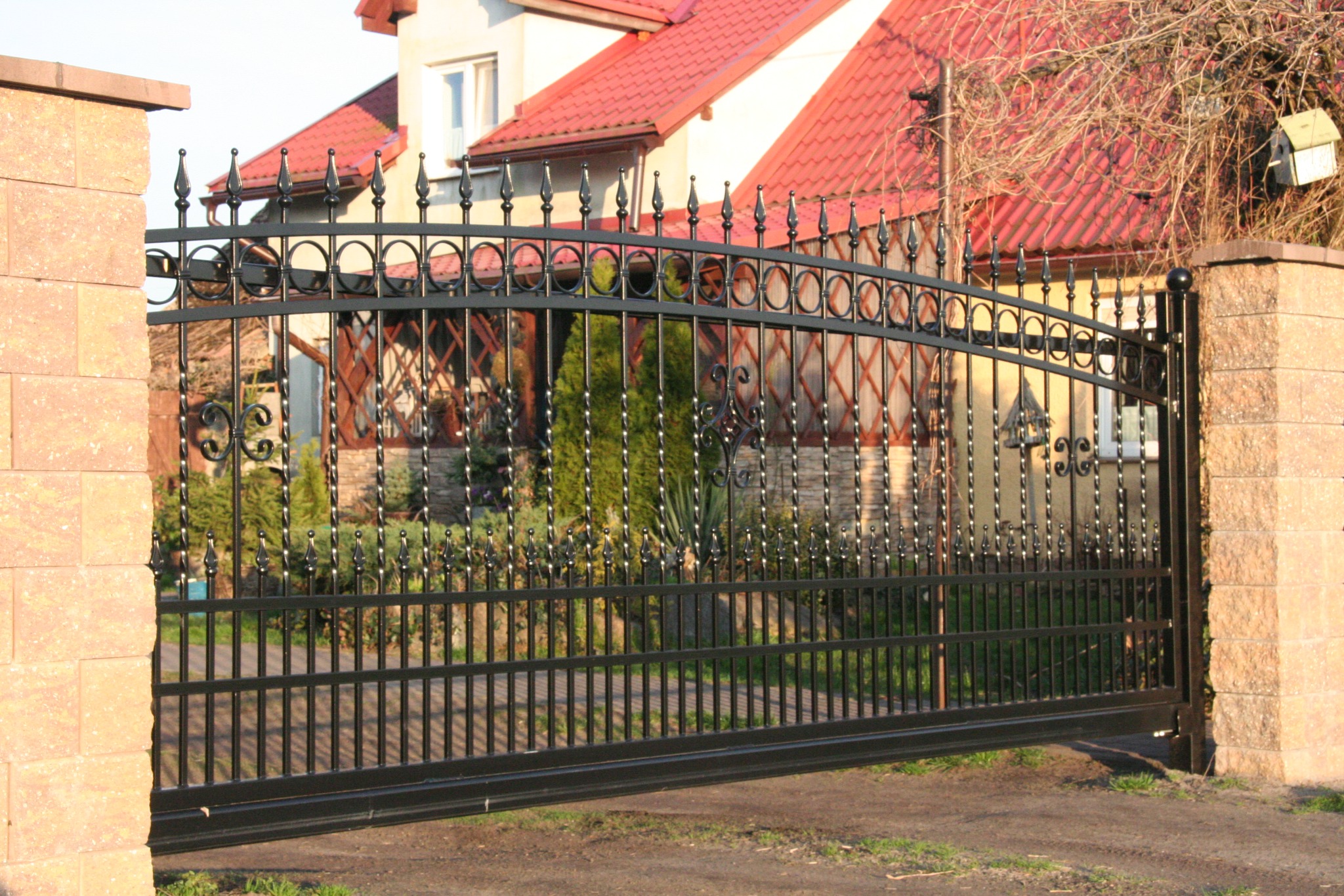 Otwarta, czarna, metalowa brama kuta z dekoracyjnymi elementami na tle domu z czerwoną dachówką i kamiennych słupków ogrodzeniowych. Widok z bliska.