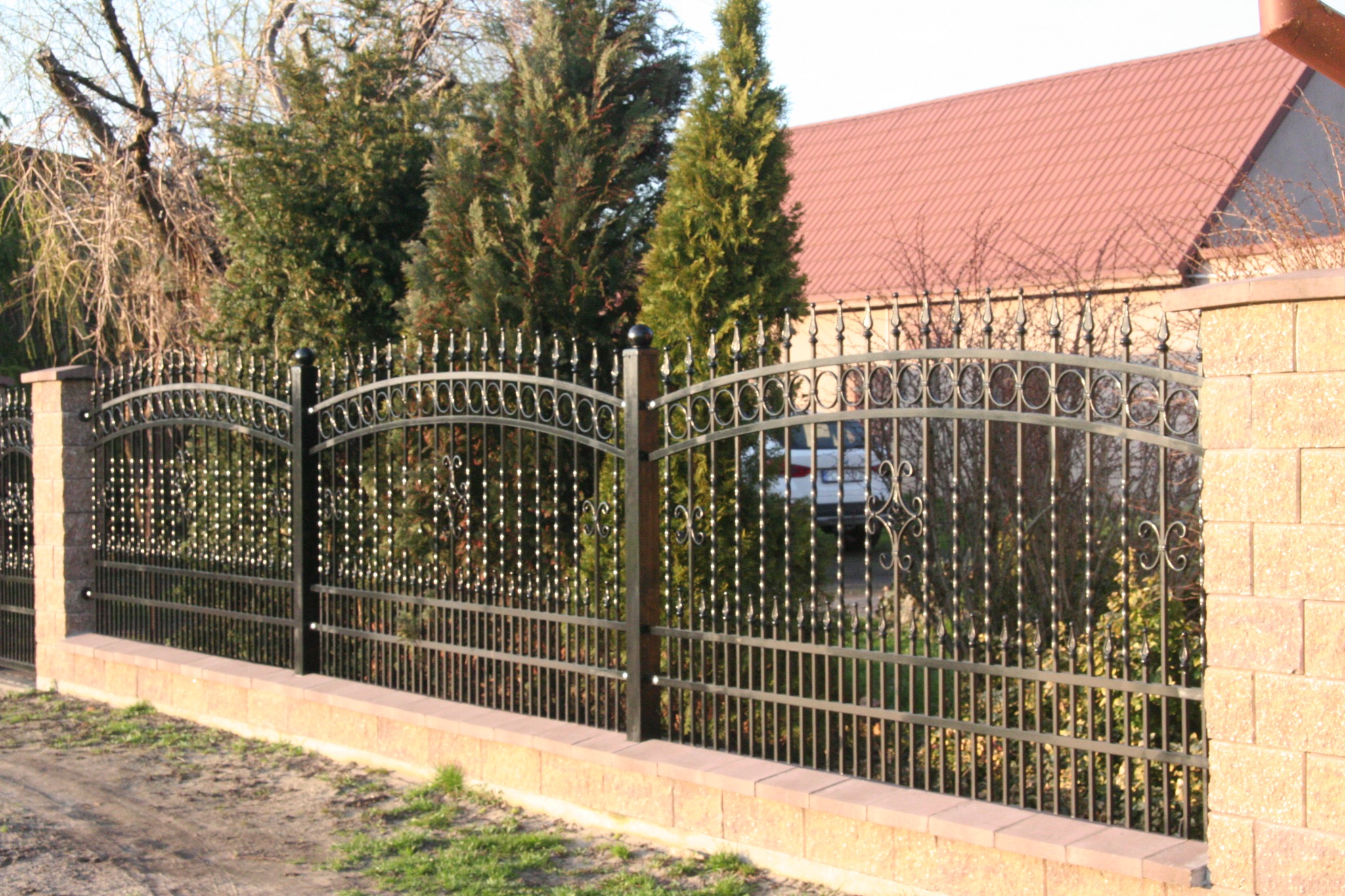 Eleganckie ogrodzenie kute z ceglanymi słupkami i podmurówką, zdobione ornamentami, na tle zieleni i domu z czerwoną dachówką. Widok na podjazd i zaparkowany samochód.
