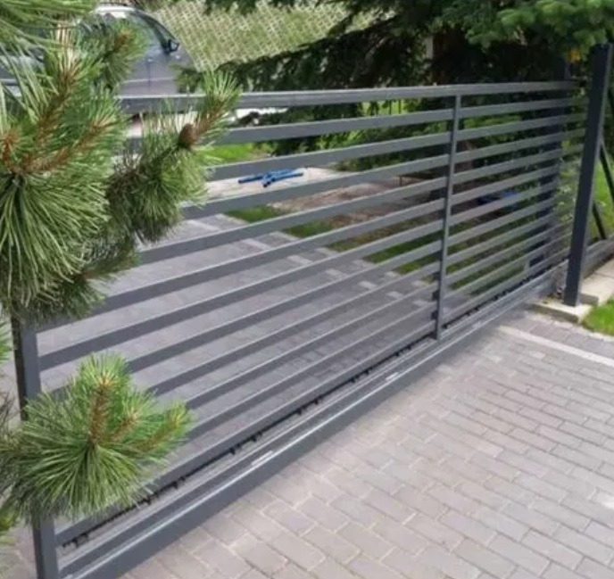 Nowoczesna, przesuwna brama wjazdowa z poziomych profili, pomalowana na szaro, z widocznym mechanizmem jezdnym. Na pierwszym planie gałązka iglaka.