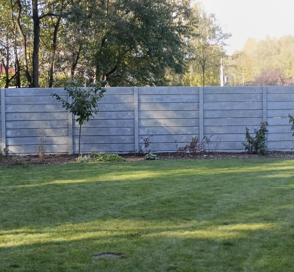 Szary, prefabrykowany płot betonowy z poziomymi panelami, oddzielający trawnik od drzew. Małe drzewko posadzone blisko ogrodzenia. Słoneczny dzień.