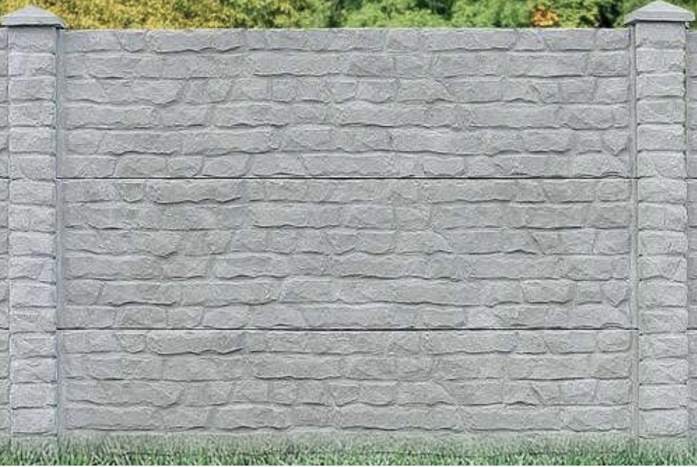 Szary, betonowy mur imitujący kamień, z wyraźną fakturą i podziałem na segmenty. Widoczne słupki z dekoracyjnymi zakończeniami. Trawa u dołu, zieleń w tle.