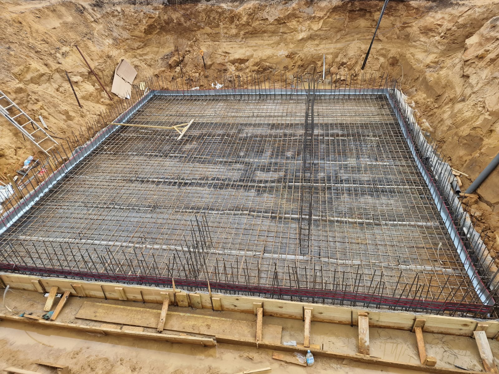 Zbrojenie fundamentu w wykopie, widok z góry. Siatka z prętów stalowych, szalunek drewniany i metalowe profile. Etap budowy domu, przygotowanie do wylania betonu.