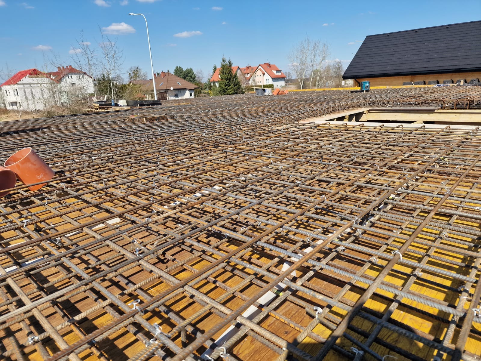Siatka zbrojeniowa na deskowaniu, przygotowana pod wylewkę fundamentów domu jednorodzinnego. W tle zabudowa mieszkalna i błękitne niebo.