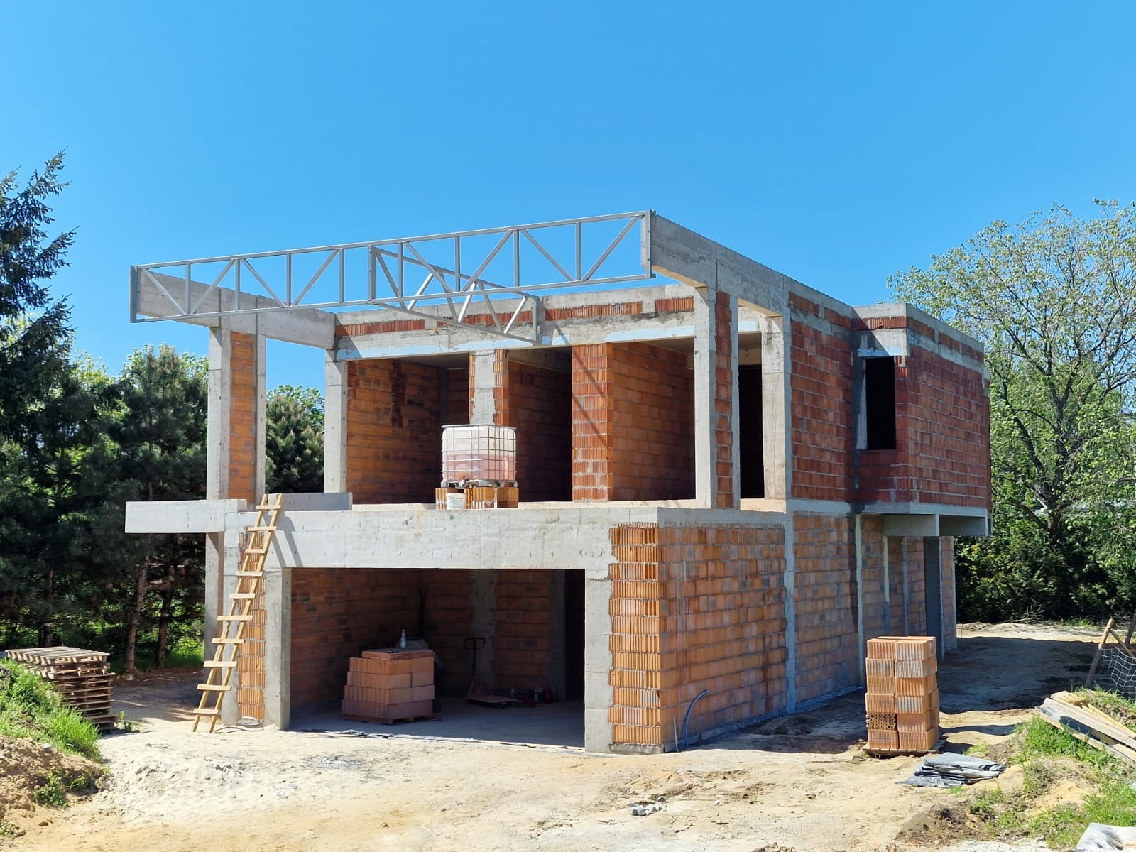 Dwukondygnacyjny dom w trakcie budowy: ściany z czerwonej cegły, betonowe słupy i strop, metalowa konstrukcja dachu. Widoczne rusztowania i materiały budowlane.