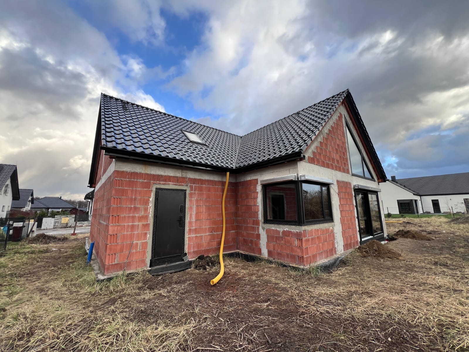 Surowy dom z czerwonej cegły z czarnym dachem i oknami, żółta rura spustowa, pochmurne niebo w tle. Widoczne detale murarskie i elementy wykończeniowe.