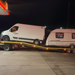 Usługi Transportowe GUTEK CAR DARIUSZ MRÓZ - Laweta z dwoma białymi busami i przyczepą kempingową na platformie, postój na stacji benzynowej nocą, widoczny pracownik w odblaskowej kamizelce.