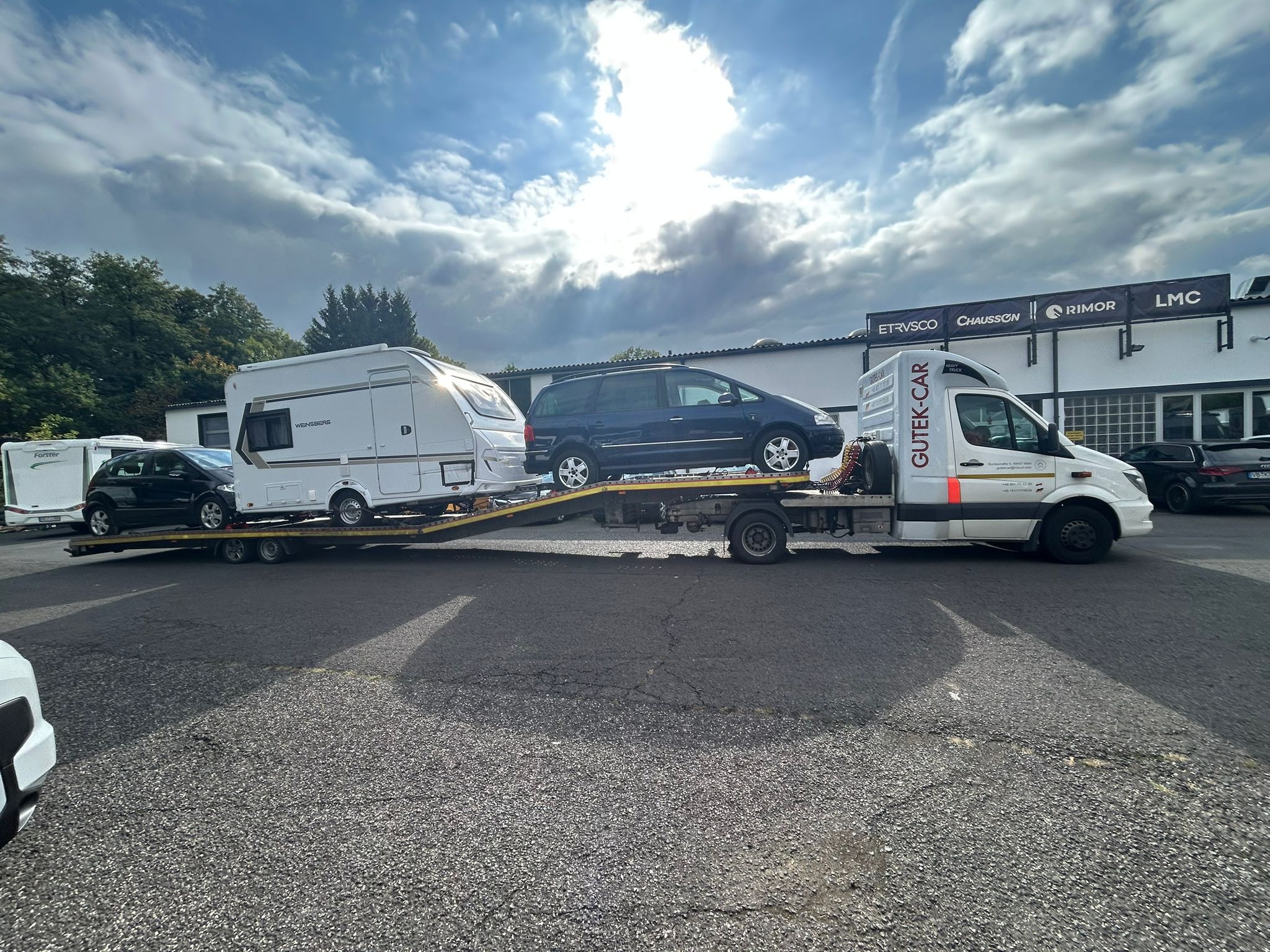 Laweta przewozi kampera i samochód osobowy na tle pochmurnego nieba. Widoczny budynek z logo producentów przyczep kempingowych.