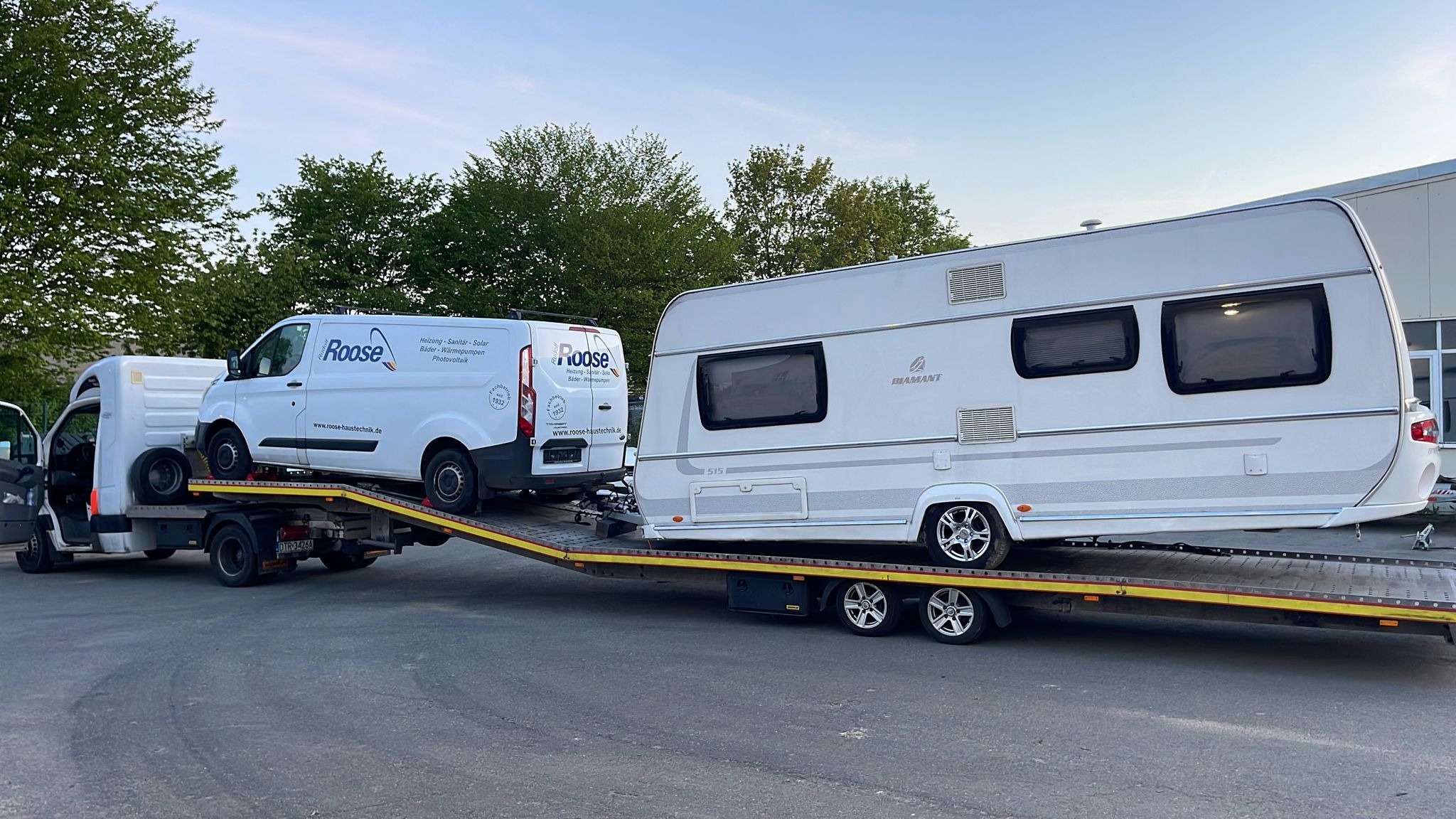 Laweta przewozi biały kamper i busa firmy Roose. Widoczne logo firmy na busie. Transport specjalistyczny na żółtej platformie. Drzewa w tle.