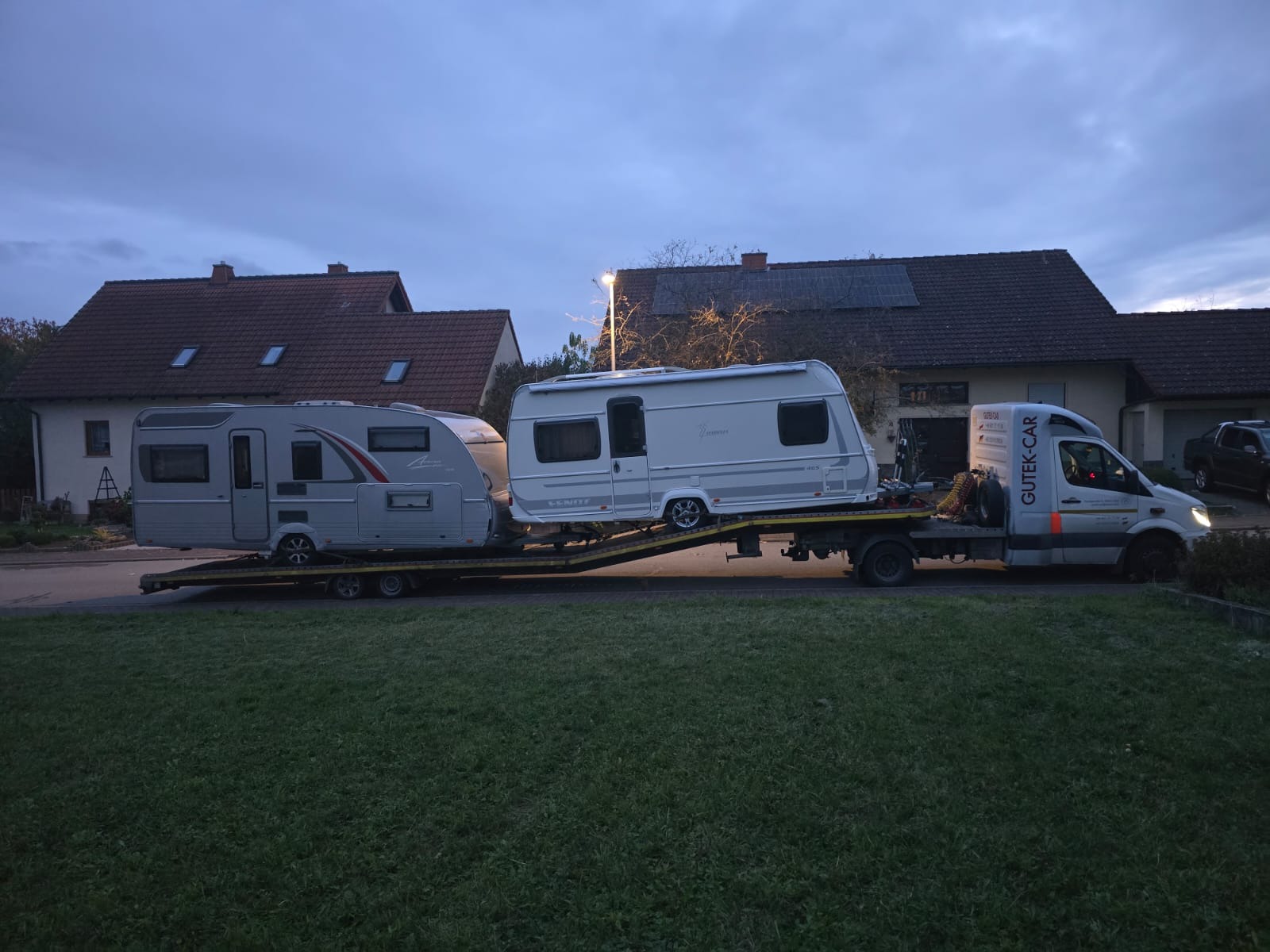 Laweta przewozi dwa kempingi na platformie transportowej. Widoczny budynek w tle i logo firmy przewozowej na pojeździe. Ujęcie z poziomu trawnika.