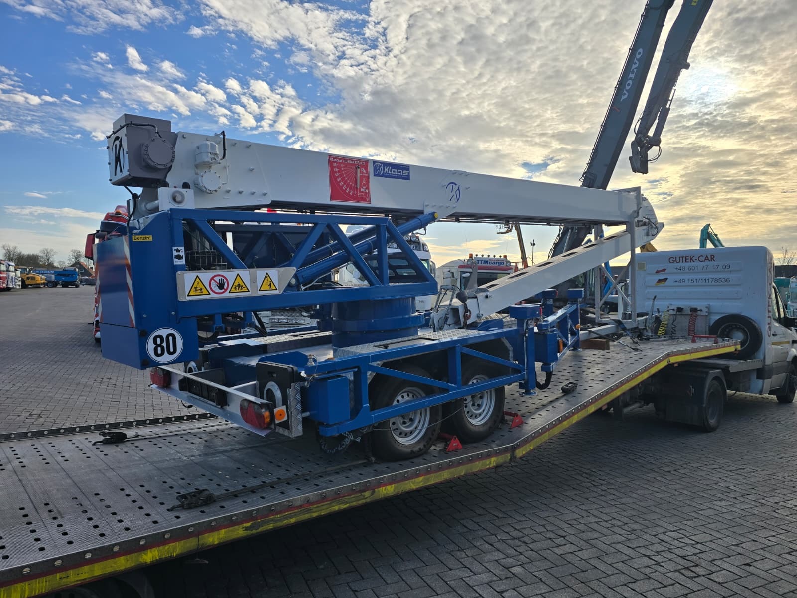 Laweta z platformą przewozi niebiesko-biały podnośnik Klaas na tle pochmurnego nieba. Widoczny numer 80 i logo Gutek-Car na pojeździe.