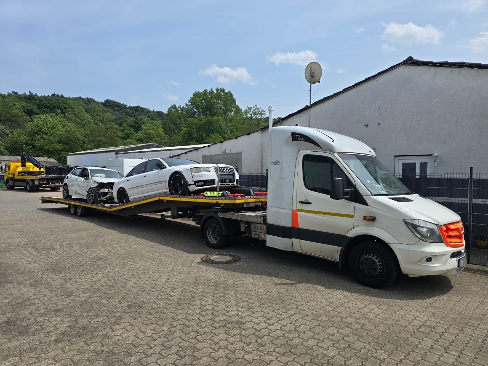 Biała laweta z dwoma uszkodzonymi, białymi samochodami osobowymi na platformie, zaparkowana na brukowanym placu, z zielenią w tle i żółtą lawetą w oddali.