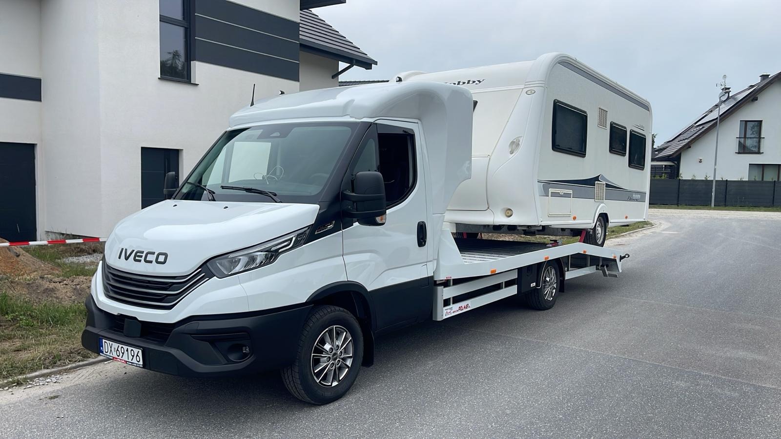 Biała laweta Iveco z przyczepą kempingową Hobby na tle nowoczesnych domów jednorodzinnych. Tablica rejestracyjna widoczna. Dzień, pochmurno.
