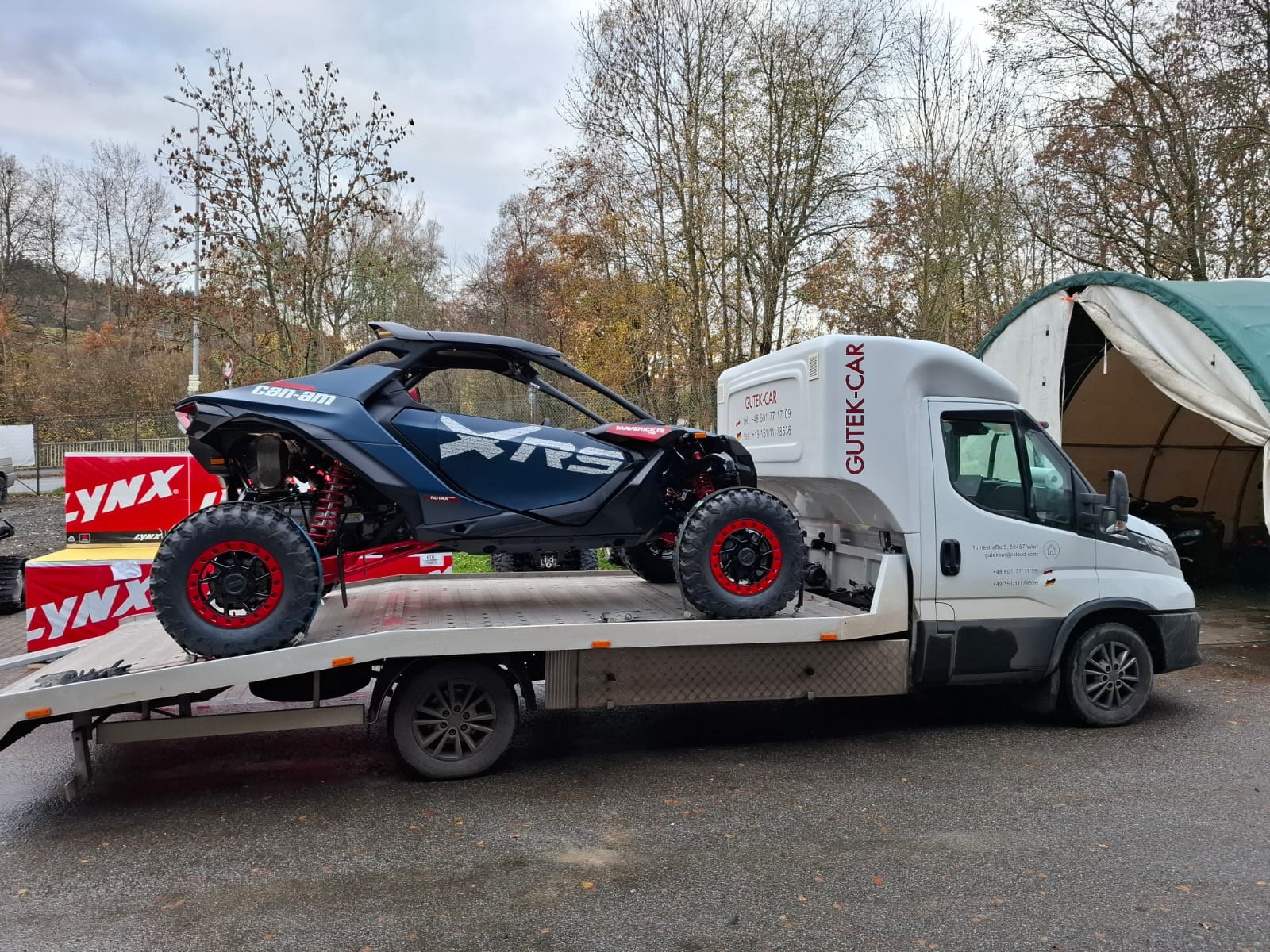 Laweta przewozi terenowy pojazd Can-Am Maverick XRS z czerwonymi felgami. Biała laweta z napisem 'GUTEK-CAR' na tle drzew i namiotu.