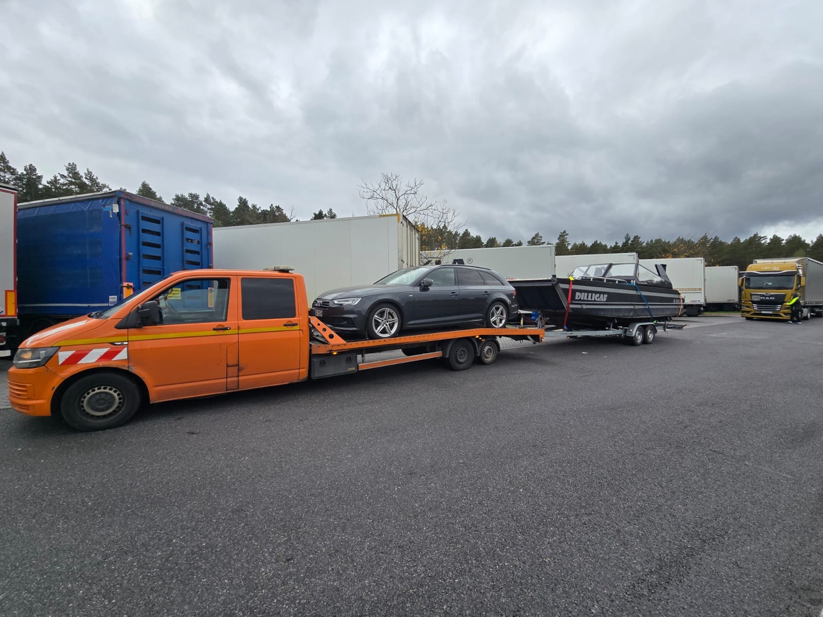 Pomarańczowa laweta z szarą osobówką i czarną łodzią na przyczepie, na tle zaparkowanych ciężarówek i pochmurnego nieba. Widok z boku, lekki kąt.