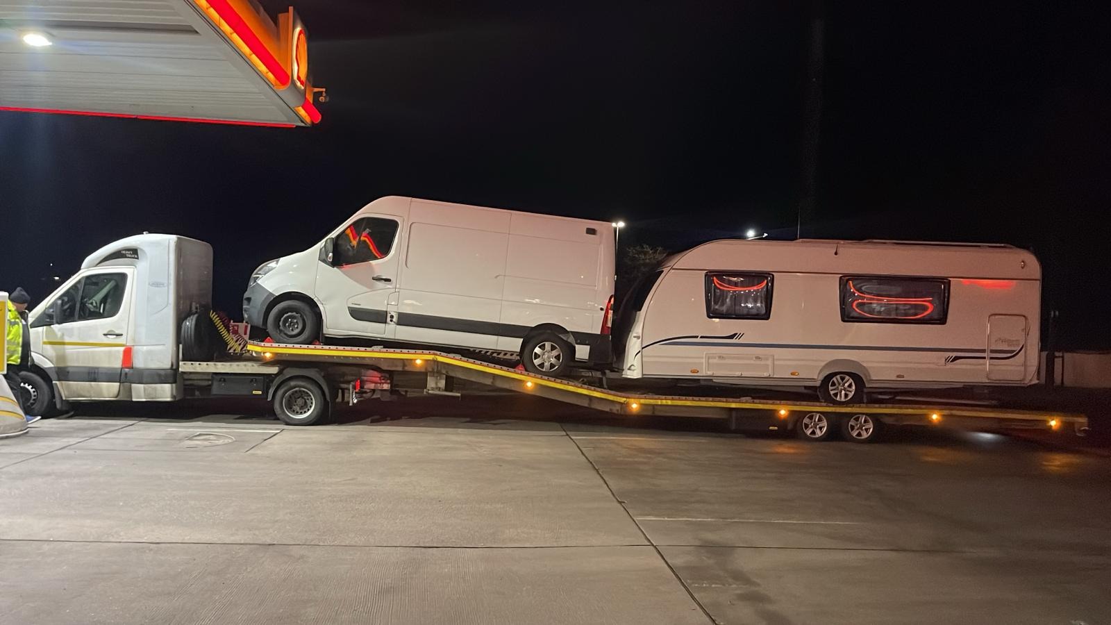 Laweta z dwoma białymi busami i przyczepą kempingową na platformie, postój na stacji benzynowej nocą, widoczny pracownik w odblaskowej kamizelce.