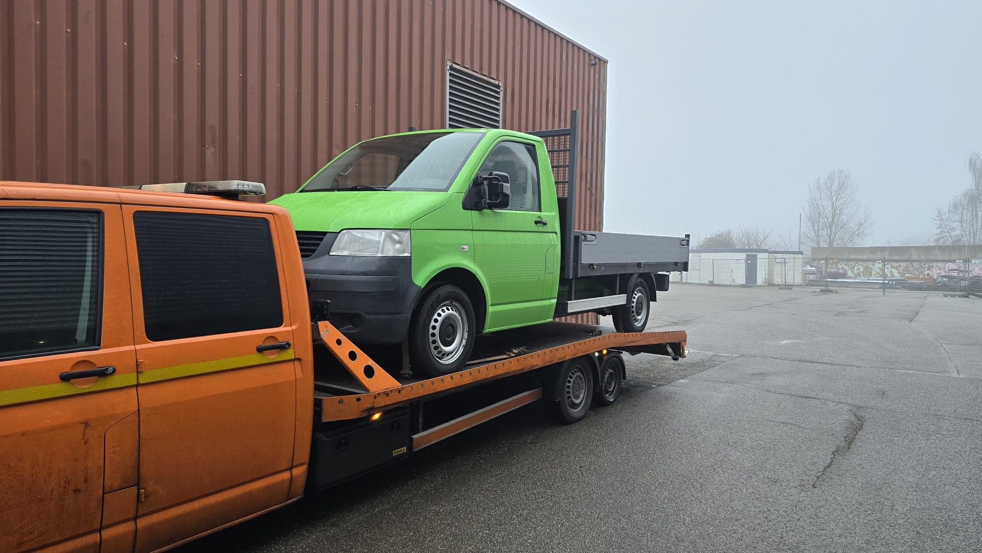 Laweta z pomarańczową kabiną transportuje zielonego busa na tle magazynu w pochmurny dzień. Widoczne detale mocowania pojazdu.
