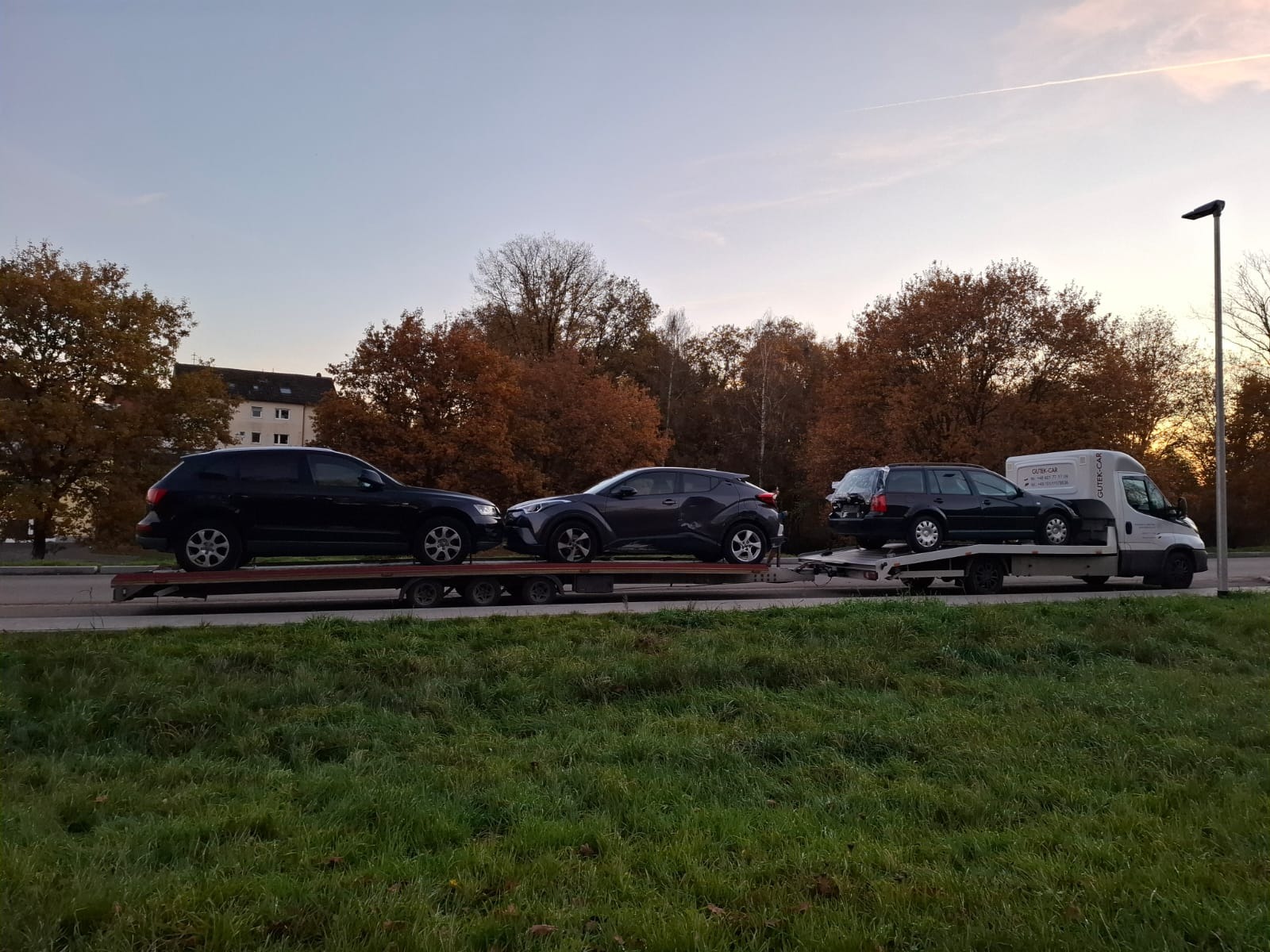 Laweta przewozi trzy samochody osobowe na tle jesiennego krajobrazu. Widoczny budynek w oddali i słup oświetleniowy. Lekko pochmurne niebo.