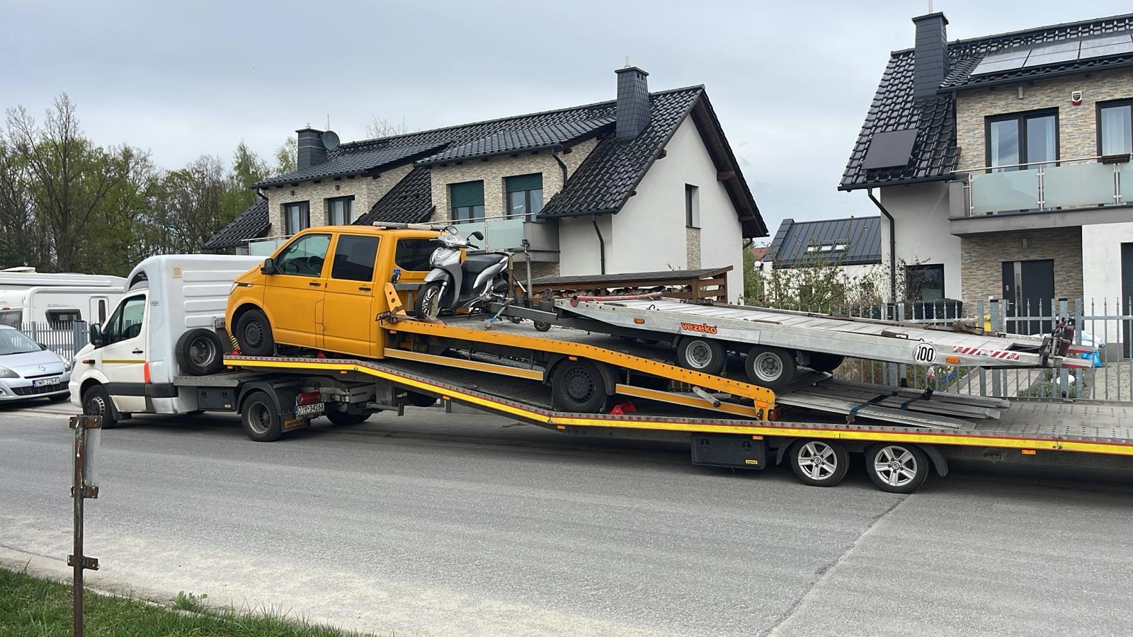 Laweta z żółtym VW Transporter i skuterem na platformie, transport drogowy na tle domów jednorodzinnych w pochmurny dzień.