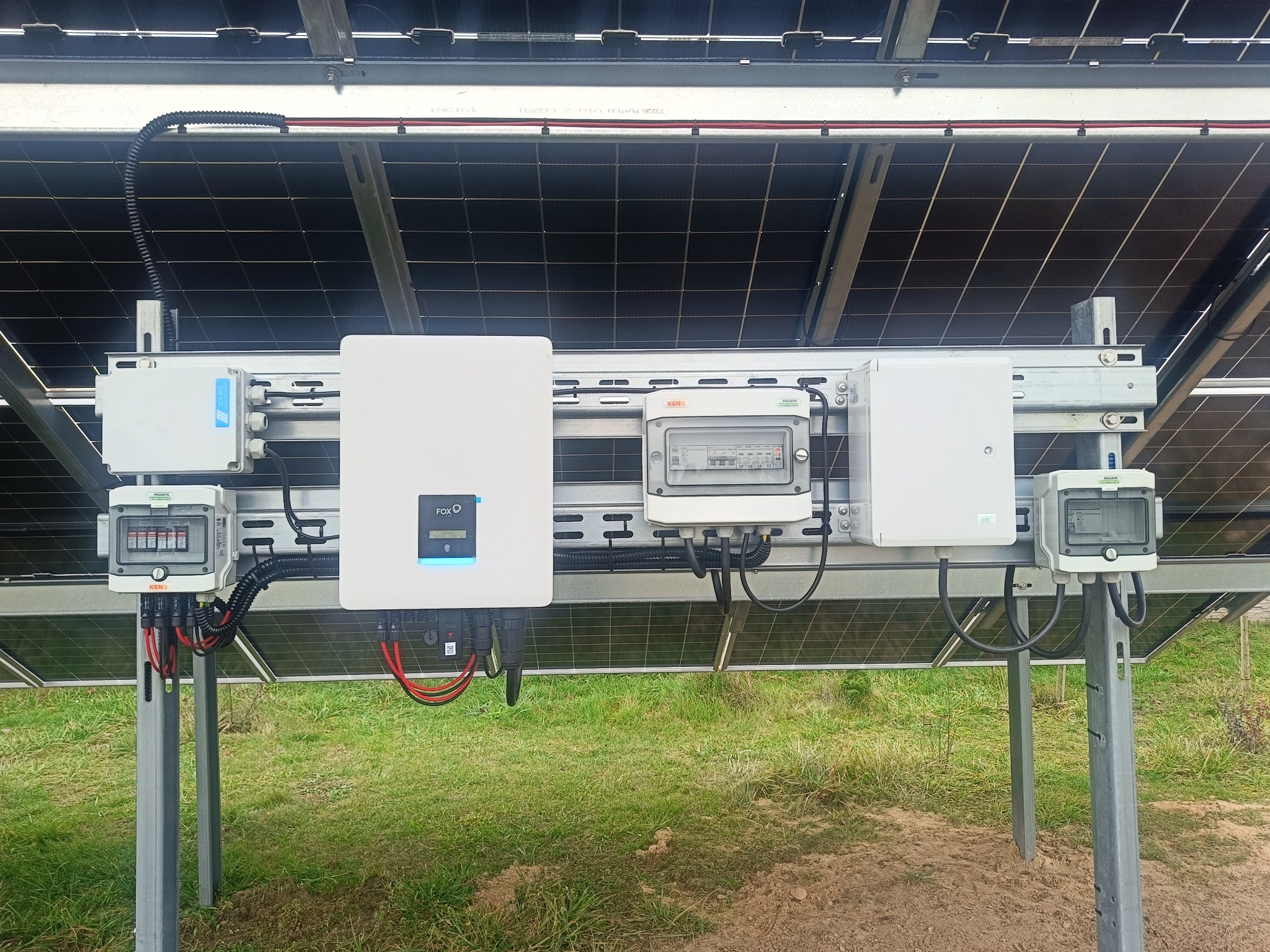 Instalacja fotowoltaiczna: widok od spodu na panele, inwerter FOX i skrzynki zabezpieczeń. Widoczne przewody i elementy montażowe na tle zielonej trawy.