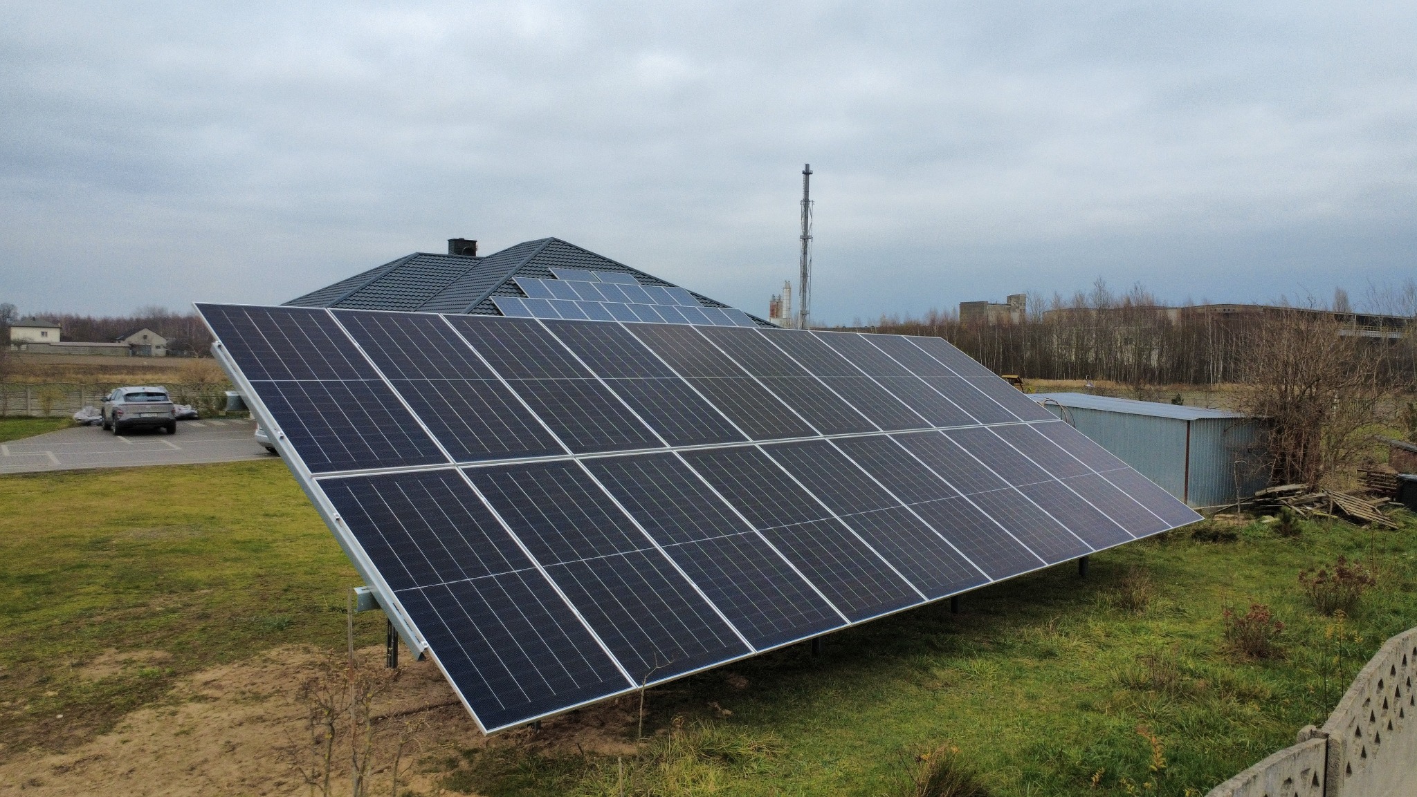 Panel fotowoltaiczny na trawie, z domem i kominem w tle. Widoczne inne panele na dachu. Pochmurne niebo. Ujęcie z góry, lekko z boku.