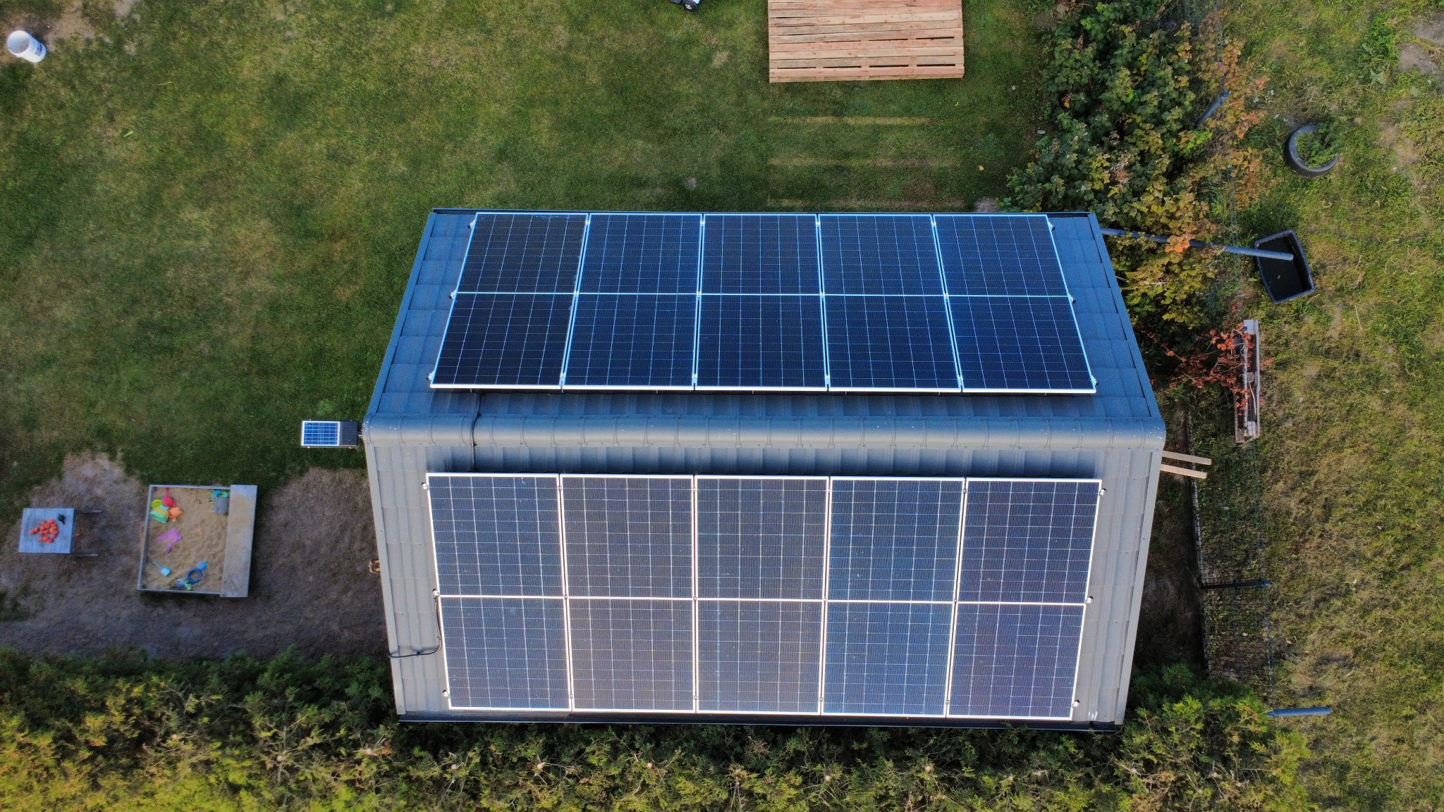 Widok z góry na dach budynku z panelami słonecznymi, obok trawnik, piaskownica i drewniane elementy. Instalacja fotowoltaiczna na dachu budynku gospodarczego.