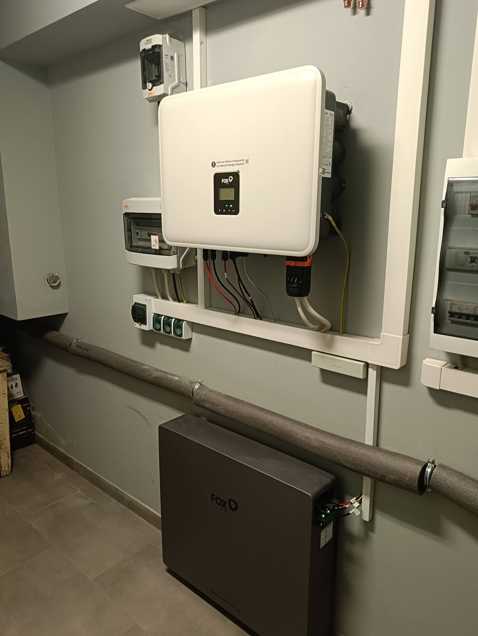 Kompletna instalacja magazynu energii Fox ESS z inwerterem i akumulatorem, zamontowana na szarej ścianie w pomieszczeniu technicznym. Widoczne przewody i zabezpieczenia.