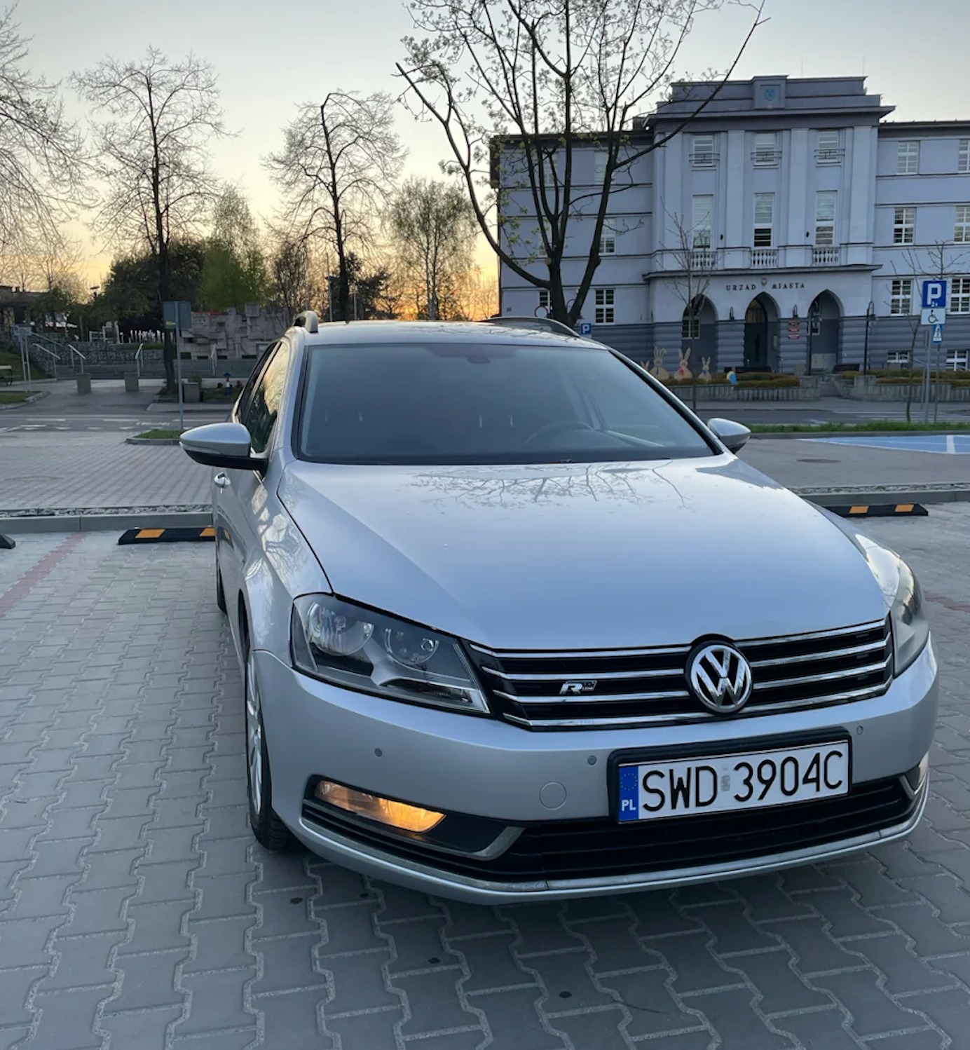 Srebrny Volkswagen Passat Variant R-Line na tle Urzędu Miasta, zaparkowany na kostce brukowej. Widoczne drzewa i zachodzące słońce.
