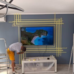 RENE KĘDROŃ - Pracownik zabezpiecza ścianę taśmą malarską wokół telewizora przed malowaniem. Widoczna drabina, stolik zabezpieczony folią. Ściana w odcieniach szarości, nowoczesne oświetlenie.