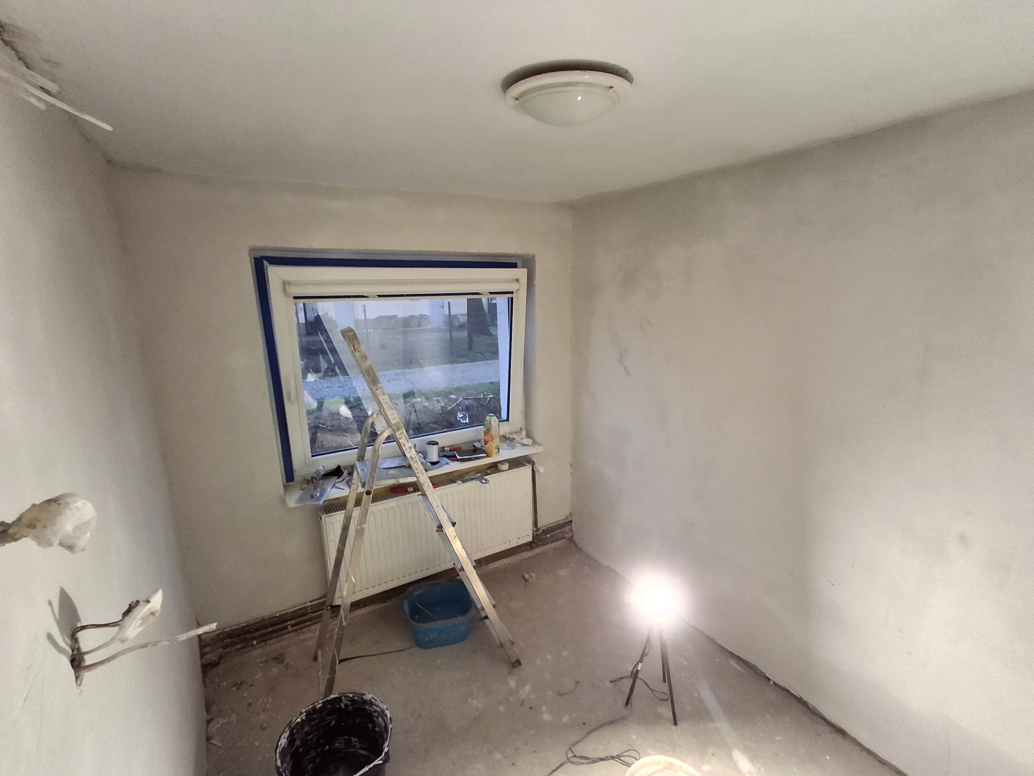 Pokój w trakcie szpachlowania: ściany pokryte gładzią, drabina oparta o okno z niebieską taśmą, widoczne narzędzia i oświetlenie budowlane. Prace wykończeniowe.