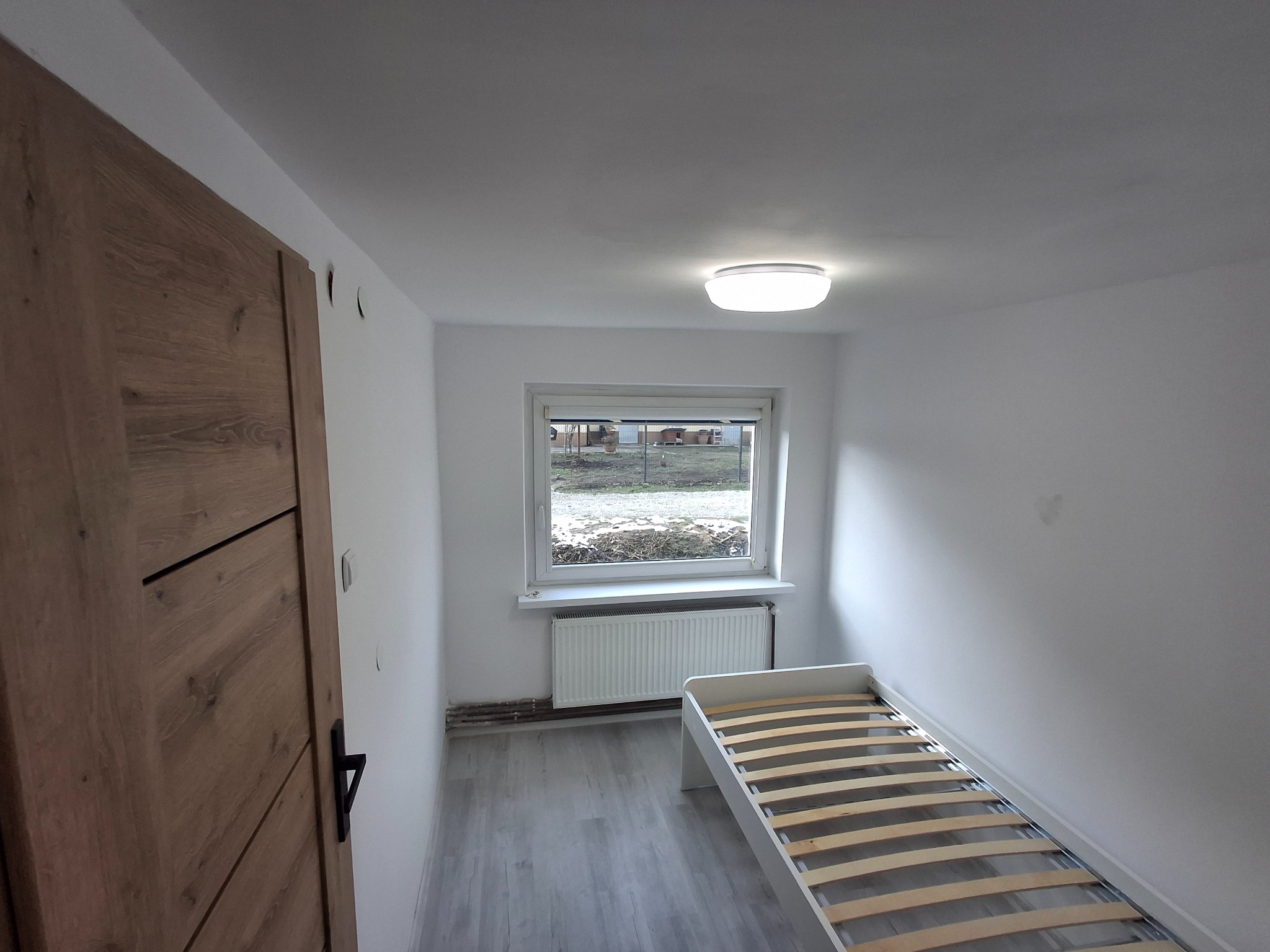 Jasne wnętrze pokoju po remoncie: białe ściany, szara podłoga, drewniane drzwi z czarną klamką, okno z grzejnikiem i częściowo złożone białe łóżko. Minimalistyczny design.