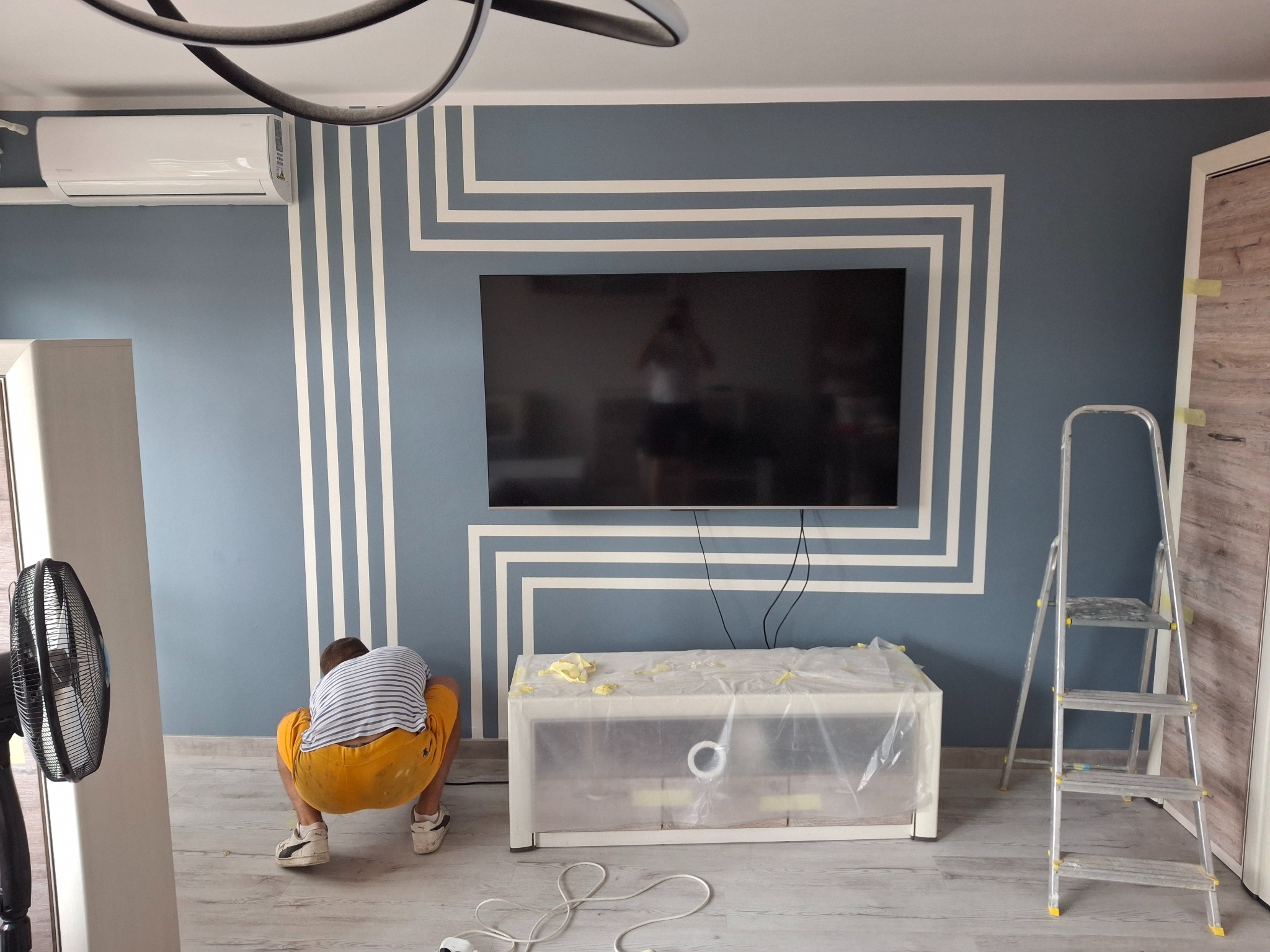 Malowanie dekoracyjne ściany w Nysie. Niebieska ściana z białymi, geometrycznymi pasami. Pracownik w trakcie pracy, zabezpieczone meble, drabina. Nowoczesny design.