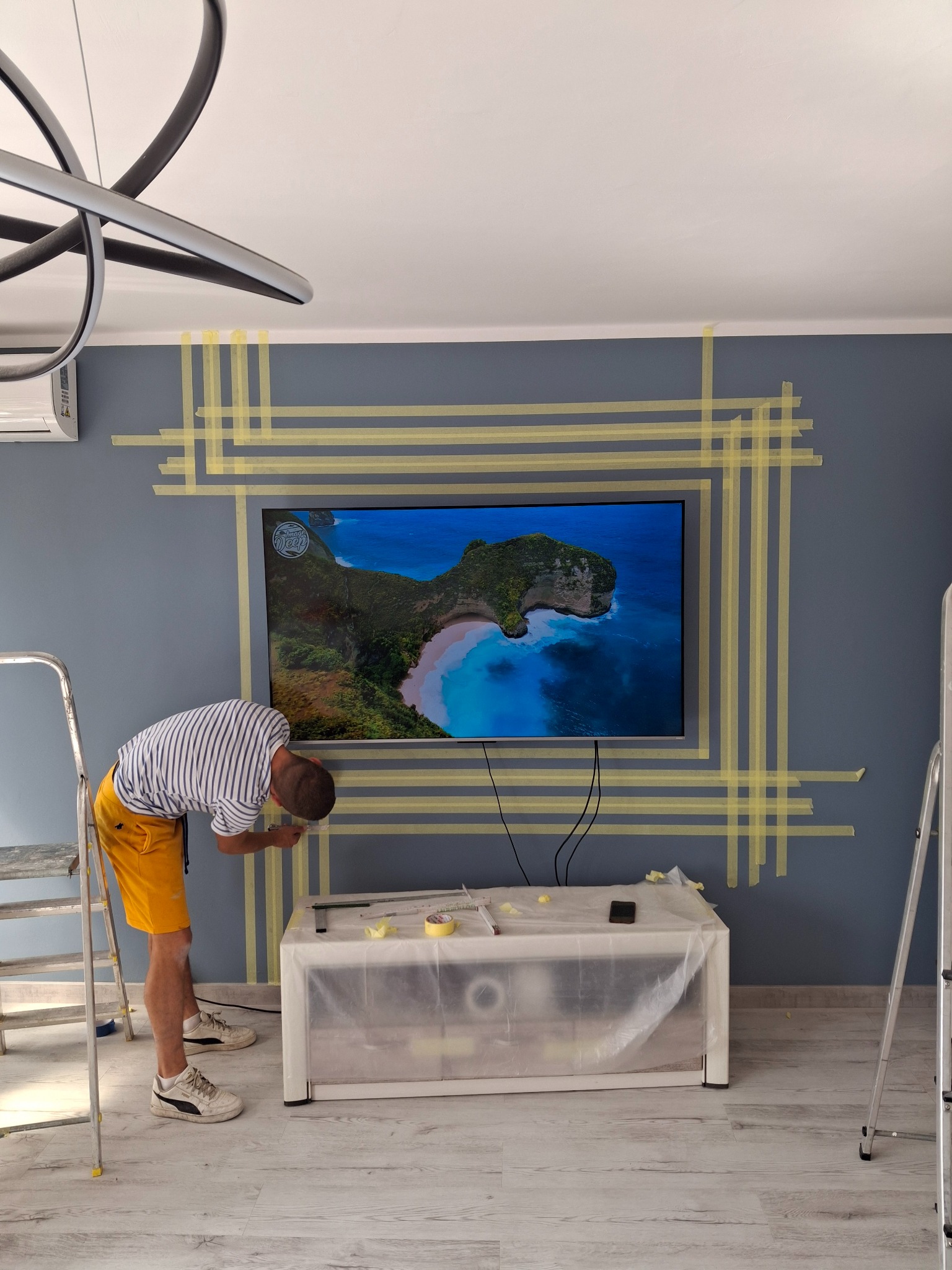 Pracownik zabezpiecza ścianę taśmą malarską wokół telewizora przed malowaniem. Widoczna drabina, stolik zabezpieczony folią. Ściana w odcieniach szarości, nowoczesne oświetlenie.
