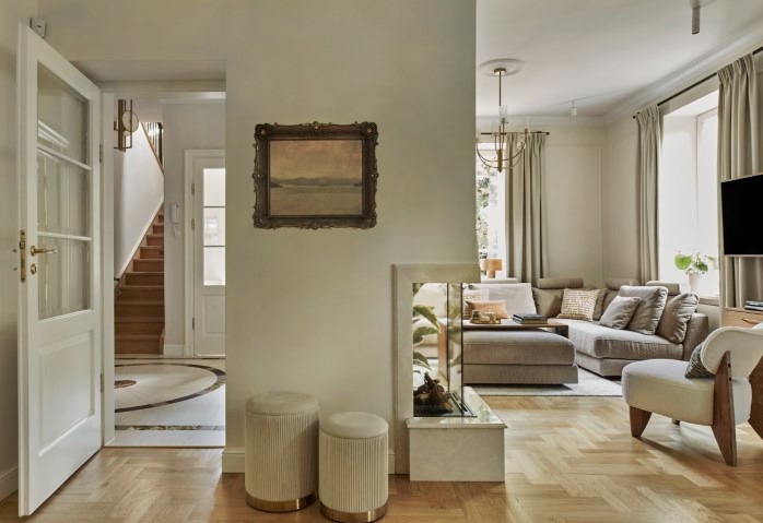 Eleganckie wnętrze willi: parkiet w jodełkę, kremowe ściany, obraz w złotej ramie, sofa narożna i dekoracyjne pufy. Widok na schody i salon.