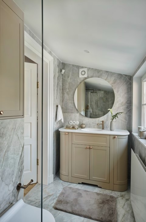 Jasna łazienka z marmurowymi płytkami, okrągłym lustrem i beżową szafką pod umywalką. Widoczny fragment kabiny prysznicowej w odbiciu lustra.