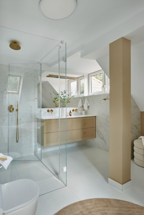 Jasna łazienka na poddaszu z kabiną prysznicową ze szkła, marmurową ścianą, drewnianą szafką pod umywalką i złotą armaturą. Widoczne okna dachowe i beżowy filar.