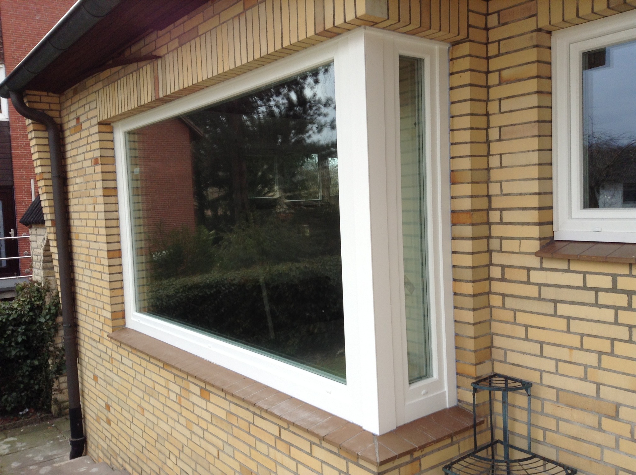 Duże, narożne okno PCV z białymi ramami, wkomponowane w elewację z żółtej cegły. Widoczne odbicie drzew w szybie.