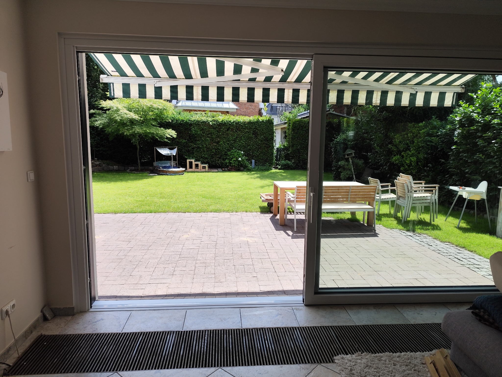 Otwarty system przesuwny z widokiem na zielony ogród i markizę w paski. Jasne płytki podłogowe i grzejnik kanałowy wewnątrz pomieszczenia.