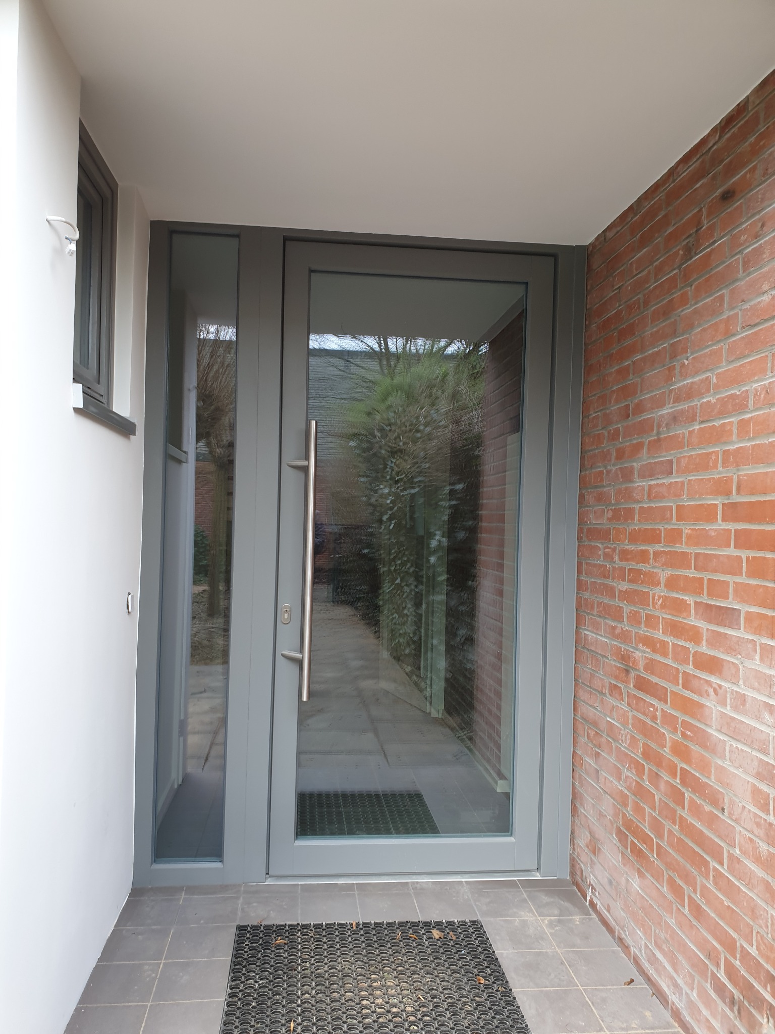 Szklane drzwi wejściowe w szarej ramie z długim, pionowym uchwytem, widoczne z zewnątrz na tle ceglanej ściany i szarej wycieraczki.
