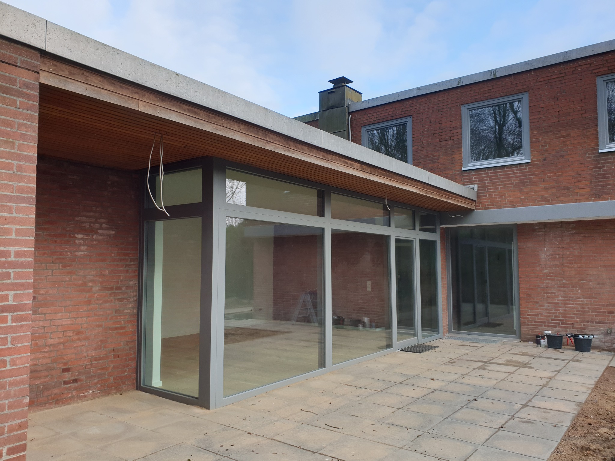 Szklana fasada budynku z cegły z szarymi ramami okiennymi i drzwiami przesuwnymi, widok z zewnątrz na taras z kostki brukowej.